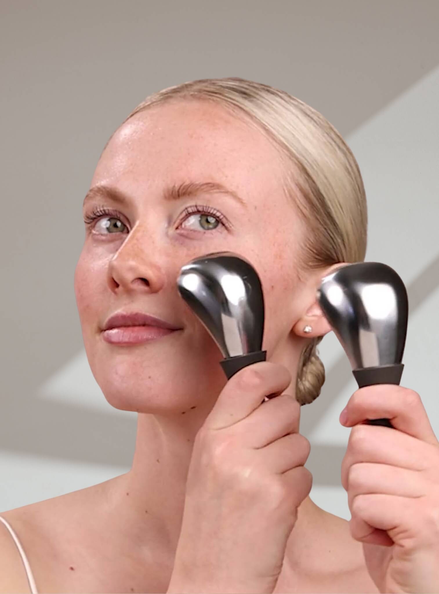 Woman using two black handheld Cryo devices on her face against a neutral background